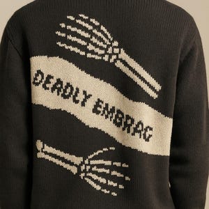 Puede incluir: Suéter de punto negro con una pancarta crema en la espalda. La pancarta dice "DEADLY EMBRAG" en letras negras, con manos esqueléticas blancas arriba y abajo. Puños y dobladillo acanalados.