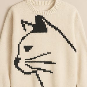 May include: Cream-colored sweater featuring a pixelated black cat face design. The crew neck sweater has long sleeves and a ribbed hem and cuffs. A simple, yet stylish, knitwear piece.