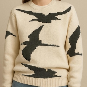 Könnte beinhalten: Cremefarbener Pullover mit einem Muster aus dunkelgrauen Vögeln im Flug. Der Rundhalspullover hat lange Ärmel und gerippte Säume. Das Design zeigt ein sich wiederholendes Muster von Vögeln auf der Vorderseite und den Ärmeln.