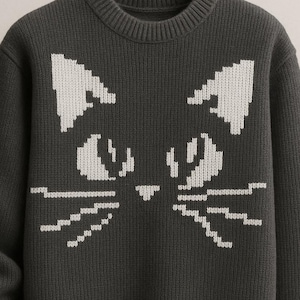 Op de afbeelding: Donkergrijze gebreide trui met een wit kattengezichtontwerp. De ogen, neus en snorharen van de kat zijn gemaakt met witte breisteken. De trui heeft een ronde hals en geribde manchetten en zoom.