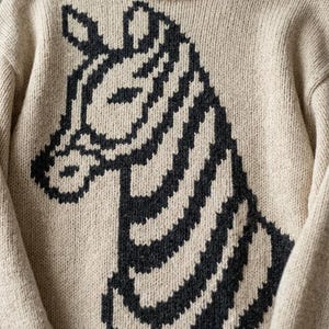 Peut inclure: Un pull en tricot beige avec un grand motif de zèbre noir sur le devant. Le pull a un col rond et des manches longues. La tête et les rayures du zèbre sont bien en évidence, créant un look graphique audacieux.