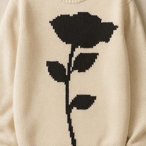 Può includere: Maglione color crema con un grande motivo floreale nero sul davanti. Il maglione girocollo ha maniche lunghe e polsini e orlo a coste. La grafica floreale comprende un fiore e uno stelo con foglie.
