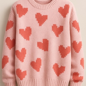 May include: A light pink sweater featuring a pattern of coral-colored pixelated hearts. The sweater has a crew neck and ribbed cuffs and hem. This knitwear is suitable for casual wear.