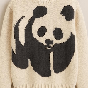 May include: Cream-colored knit sweater featuring a large black panda bear graphic on the front. The sweater has a crew neck and ribbed cuffs and hem. A cozy and casual top for everyday wear.