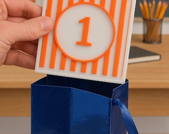 May include: A white and orange sign with the text "Whatateacher" and the number "1" is held above a blue gift bag. The sign has orange vertical stripes and a circular design around the number. The gift bag has a shiny finish.
