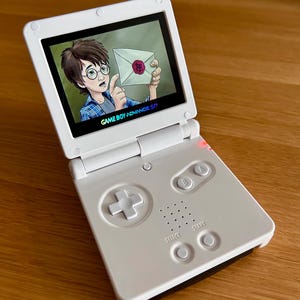 Könnte beinhalten: Ein weißes Game Boy Advance SP-Gerät, das ein farbenfrohes Bild eines Zeichentrickcharakters mit einem Brief zeigt. Der Bildschirm zeigt den Text "GAME BOY ADVANCE SP". Die Konsole verfügt über ein Steuerkreuz, Tasten und Start/Select-Tasten.
