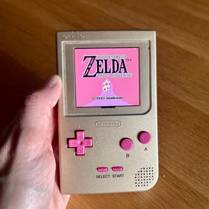 Könnte beinhalten: Ein goldfarbener Handheld-Spielautomat mit einem rosa Bildschirm, auf dem "The Legend of Zelda: Link's Awakening" angezeigt wird. Das Gerät hat rosa Knöpfe und das Nintendo-Logo. Das Spiel wurde 1993 veröffentlicht.