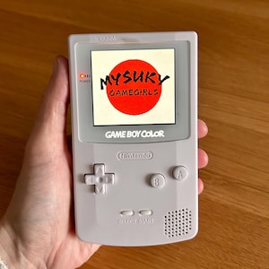 Może przedstawiać: Jasnoszara konsola przenośna Game Boy Color z tekstem "MYSÜKY GAMEGIRLS" w czerwonym okręgu na ekranie. Posiada krzyżak, przyciski A i B oraz przyciski start/select. Kontrolka zasilania jest włączona.