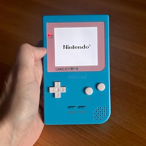 Op de afbeelding: Een turquoise Game Boy handheld gameconsole met een roze rand rond het scherm. Het scherm toont het Nintendo-logo. De console heeft een D-pad, knoppen en de tekst "GAME BOY".
