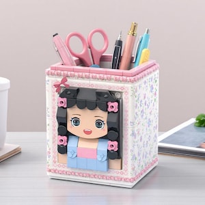 May include: A decorative desk organizer featuring a cartoon girl design. The white box is adorned with a floral pattern and pink trim. It holds pink scissors, pens, and a pencil. The girl's face is made of blocks, with black hair and a blue dress.