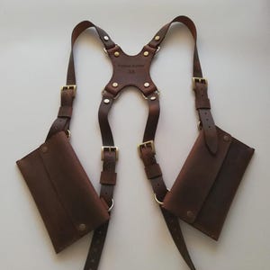 May include: A brown leather shoulder holster with two pouches. The holster features adjustable straps, brass-colored buckles, and rivets. The central piece is embossed with text and the number 33.