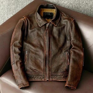 Puede incluir: Chaqueta de cuero marrón con acabado envejecido. La chaqueta tiene cremallera frontal, cuello y múltiples bolsillos. La chaqueta se muestra sobre una superficie de cuero marrón.