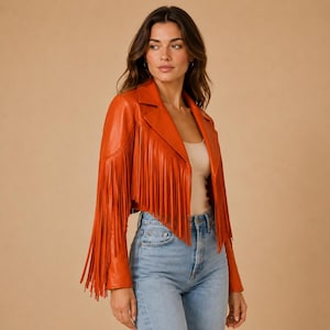 May include: A vibrant orange leather jacket featuring long fringe detailing along the front panels and sleeves. The jacket has a classic collar, a button-front closure, and a cropped silhouette, worn over a plain light-colored top and blue jeans.