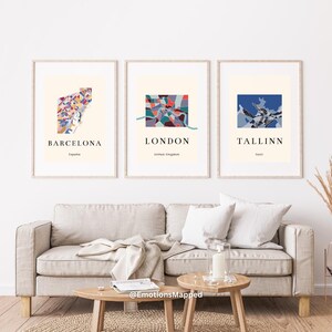 Może przedstawiać: Trzy oprawione grafiki przedstawiające kolorowe, abstrakcyjne mapy miast Barcelony, Londynu i Tallinna. Każda grafika zawiera nazwę miasta i kraju poniżej mapy. Grafiki są eksponowane nad beżową sofą z drewnianymi stolikami bocznymi.
