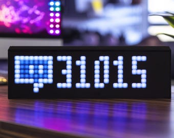 Puede incluir: Una pantalla digital negra que muestra un icono de burbuja de diálogo azul y el número 31015 en blanco, con fuente pixelada. La pantalla está sobre una superficie de madera con un fondo borroso.