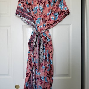May include: A pink floral print maxi dress with a blue and white patterned sash. The dress has a V-neckline and short sleeves.