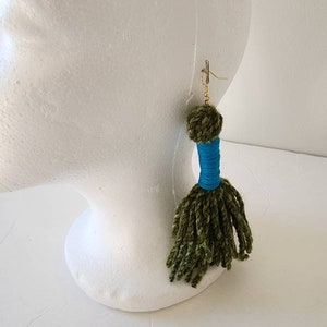 May include: Green and blue yarn tassel earrings with a gold hook. The tassel is made of a fluffy green yarn with a blue yarn band in the middle.