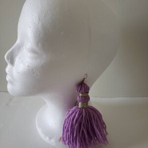 May include: Purple yarn tassel earrings with gold and silver accents. The earrings are hanging from a white mannequin head.
