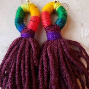 May include: A pair of rainbow-colored yarn earrings with burgundy tassels. The earrings are made with a thick yarn and have a circular shape. The tassels are long and flowy.