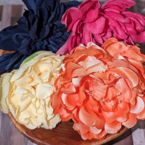 May include: Four silk flower heads in a wooden bowl. The flowers are a deep blue, a bright pink, a pale yellow, and a coral orange.