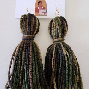 May include: A pair of handmade green, brown, and black yarn tassel earrings with gold hooks. The earrings are displayed on a white surface with a business card that reads "Stitch.Niche - handcrafted".