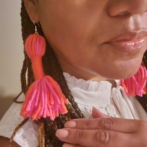May include: A pair of pink and orange tassel earrings. The earrings are long and dangle from the earlobes. The tassels are made of yarn and are very fluffy.