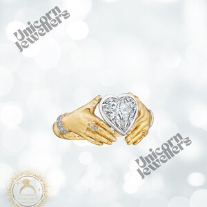 K&ouml;nnte beinhalten: Ein goldener Ring mit einem herzf&ouml;rmigen Diamanten, der von zwei H&auml;nden gehalten wird. Der Ring hat kleine Diamanten am Band und an den H&auml;nden. Der Hintergrund ist ein weiches, verschwommenes Wei&szlig; mit dem Text "Unicorn Jewellers".