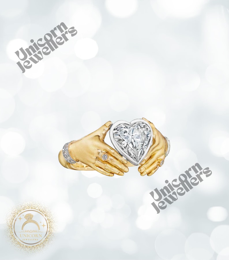 K&ouml;nnte beinhalten: Ein goldener Ring mit einem herzf&ouml;rmigen Diamanten, der von zwei H&auml;nden gehalten wird. Der Ring hat kleine Diamanten am Band und an den H&auml;nden. Der Hintergrund ist ein weiches, verschwommenes Wei&szlig; mit dem Text "Unicorn Jewellers".