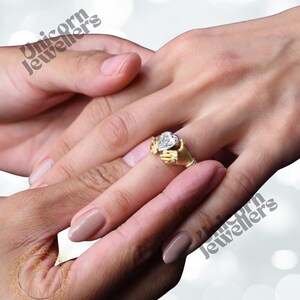 K&ouml;nnte beinhalten: Ein goldener Ring mit einem herzf&ouml;rmigen Diamanten, der von zwei kleinen H&auml;nden gehalten wird. Der Ring befindet sich an einem Finger, eine andere Hand ber&uuml;hrt ihn sanft. Der Ring ist ein Symbol f&uuml;r Liebe und Engagement.