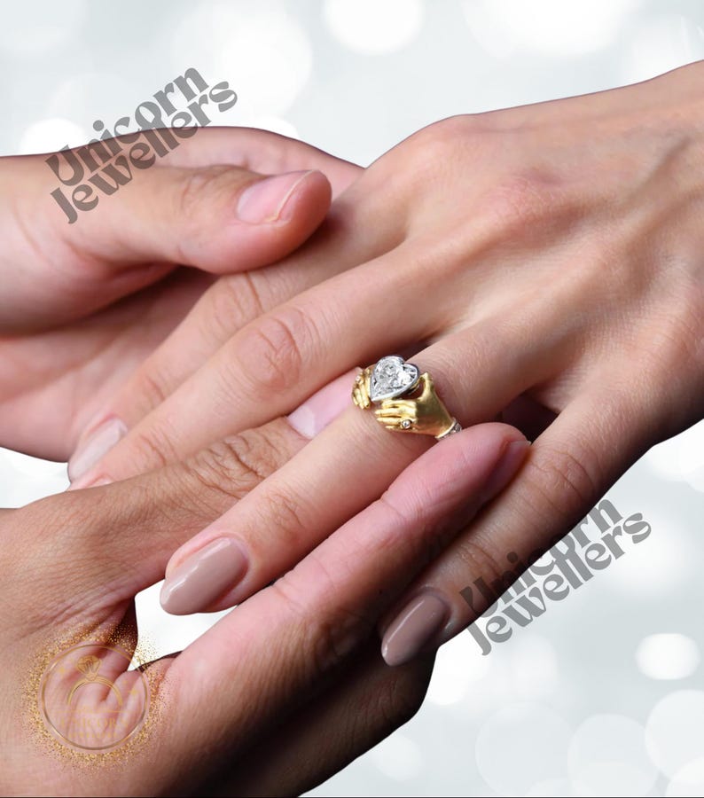 K&ouml;nnte beinhalten: Ein goldener Ring mit einem herzf&ouml;rmigen Diamanten, der von zwei kleinen H&auml;nden gehalten wird. Der Ring befindet sich an einem Finger, eine andere Hand ber&uuml;hrt ihn sanft. Der Ring ist ein Symbol f&uuml;r Liebe und Engagement.