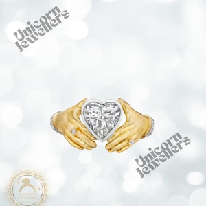 K&ouml;nnte beinhalten: Ein herzf&ouml;rmiger Diamant, der von zwei goldfarbenen H&auml;nden gehalten wird, mit Ringen an den Fingern. Der Diamant ist in einen wei&szlig;en Metallrahmen gefasst. Das Bild hat den Text "Unicorn Jewellers".