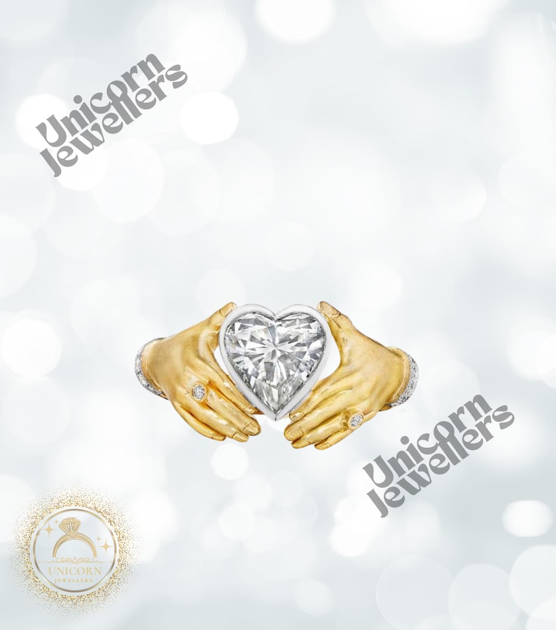 K&ouml;nnte beinhalten: Ein herzf&ouml;rmiger Diamant, der von zwei goldfarbenen H&auml;nden gehalten wird, mit Ringen an den Fingern. Der Diamant ist in einen wei&szlig;en Metallrahmen gefasst. Das Bild hat den Text "Unicorn Jewellers".