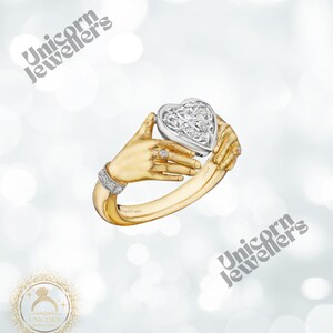 K&ouml;nnte beinhalten: Ein goldener Ring mit einem herzf&ouml;rmigen Diamanten, der von zwei stilisierten H&auml;nden gehalten wird. Der Ring hat ein diamantbesetztes Band und einen kleineren Diamantring an einem Finger. Der Text "Unicorn Jewellers" ist im Bild enthalten.