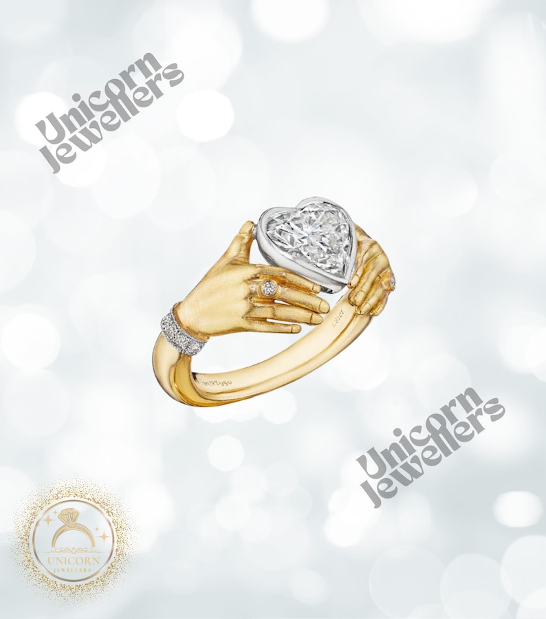 K&ouml;nnte beinhalten: Ein goldener Ring mit einem herzf&ouml;rmigen Diamanten, der von zwei stilisierten H&auml;nden gehalten wird. Der Ring hat ein diamantbesetztes Band und einen kleineren Diamantring an einem Finger. Der Text "Unicorn Jewellers" ist im Bild enthalten.