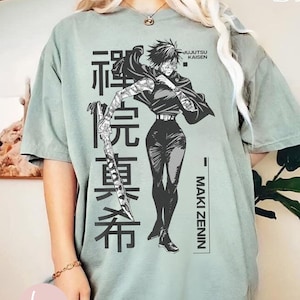 May include: Sage green t-shirt featuring a black and white illustration of Maki Zenin from Jujutsu Kaisen. The design includes Japanese text and the character's name. The shirt has short sleeves and a relaxed fit.