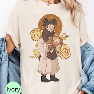 May include: Ivory-colored t-shirt featuring an anime-style illustration. The design depicts a character holding a small animal, set against a backdrop of a golden moon and clouds. The character is wearing a brown scarf and traditional clothing. The word "妹" is also visible.