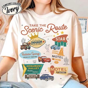 May include: Ivory Comfort Colors t-shirt with a graphic featuring the text "Take the Scenic Route" and illustrations of vintage motel signs, cars, and cacti. The design includes the text "Radiator Springs" and "Cozy Cone Motel".