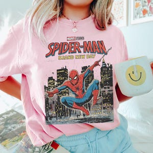 May include: A light pink sweatshirt featuring a Marvel Studios Spider-Man graphic. The graphic includes the words "Spider-Man Brand New Day" and an illustration of Spider-Man in action against a city skyline. A white mug with a yellow smiley face is also visible.