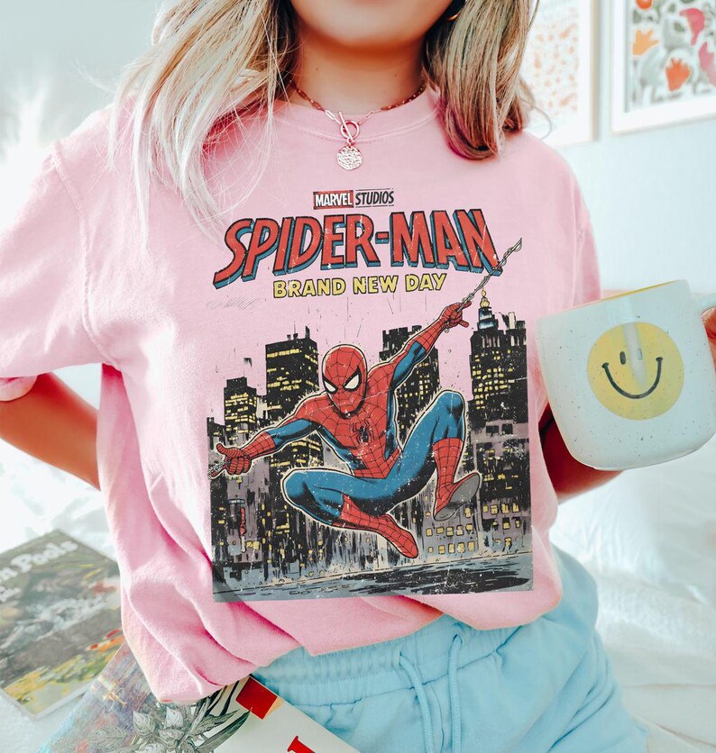 May include: A light pink sweatshirt featuring a Marvel Studios Spider-Man graphic. The graphic includes the words "Spider-Man Brand New Day" and an illustration of Spider-Man in action against a city skyline. A white mug with a yellow smiley face is also visible.