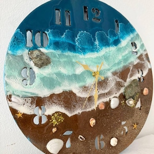 May include: A round wall clock with a beach-themed design. The top portion is a deep blue, transitioning to turquoise and white to mimic ocean waves. The bottom section is brown, resembling sand, with embedded seashells and rocks. Gold clock hands.