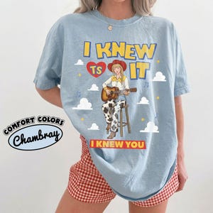May include: A light blue t-shirt featuring a graphic of a person in a cowboy hat and cow-print pants playing an acoustic guitar while sitting on a stool. The text reads I KNEW TS IT and I KNEW YOU, surrounded by clouds, stars, and musical notes.