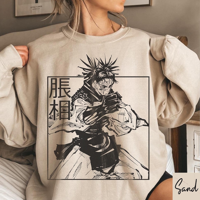 May include: Sand-colored sweatshirt featuring a black and white illustration of a character with spiky hair and a determined expression. The image is framed by a black rectangle with Japanese text. The sweatshirt has long sleeves.