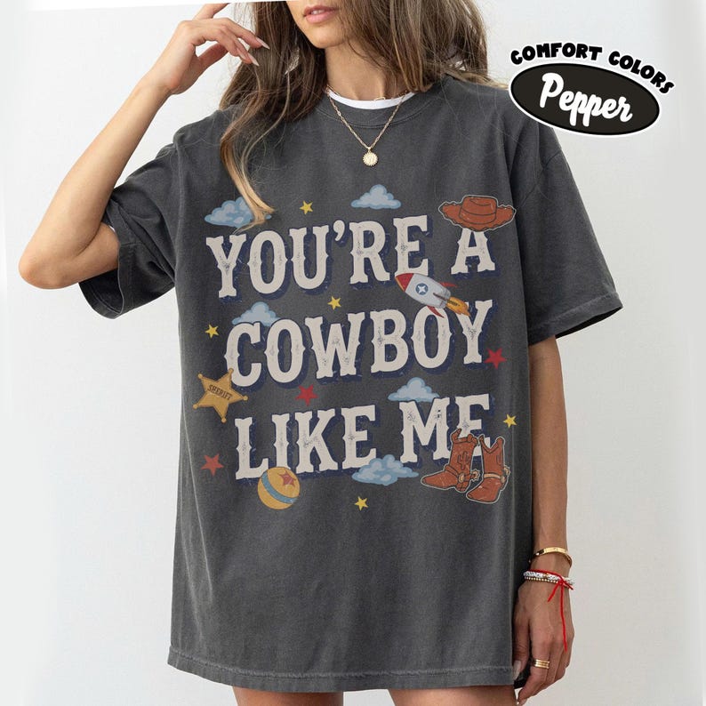 May include: A charcoal gray short-sleeve t-shirt featuring the text "YOU'RE A COWBOY LIKE ME" in white. The design includes illustrations of a brown cowboy hat, a rocket ship, a yellow sheriff star, a colorful ball, and small blue clouds and stars.