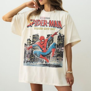 May include: Cream-colored t-shirt featuring a graphic of Spider-Man in a dynamic pose against a cityscape backdrop. The shirt includes the text "Marvel Studios Spider-Man Brand New Day" in a comic book style.