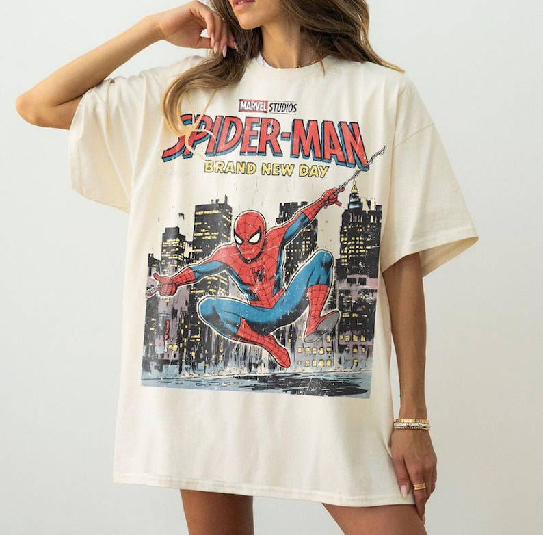 May include: Cream-colored t-shirt featuring a graphic of Spider-Man in a dynamic pose against a cityscape backdrop. The shirt includes the text "Marvel Studios Spider-Man Brand New Day" in a comic book style.