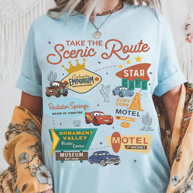 May include: Pale blue t-shirt with a graphic design featuring the text "Take the Scenic Route." The design includes car illustrations, cacti, and signs for a cafe, motel, and visitor centre. The shirt has rolled-up sleeves.