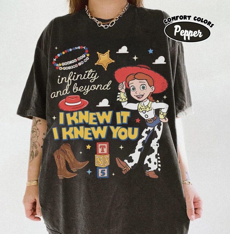May include: A charcoal gray short-sleeve t-shirt featuring a graphic print of a cowgirl character, a red cowboy hat, brown cowboy boots, and alphabet blocks. The text reads "infinity and beyond", "I KNEW IT I KNEW YOU", and "YOU'VE GOT A FRIEND IN ME".