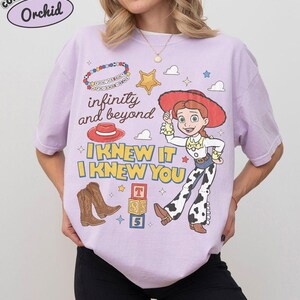 May include: A light purple t-shirt featuring a graphic print of a cowgirl character, a red hat, brown cowboy boots, a gold star, alphabet blocks, and a beaded bracelet. The text reads infinity and beyond, I KNEW IT, I KNEW YOU, and You've got a friend in me.