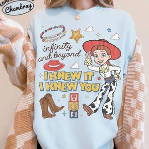 May include: A light blue crewneck sweatshirt featuring a graphic print of Jessie from Toy Story, a red cowboy hat, brown boots, a beaded bracelet, and alphabet blocks. The text reads infinity and beyond, I KNEW IT, I KNEW YOU, and YOU'VE GOT A FRIEND IN ME.