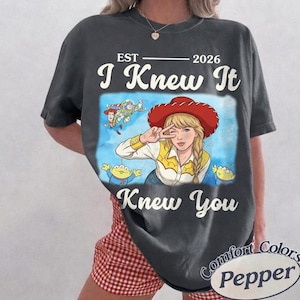 May include: A charcoal gray t-shirt featuring a graphic print of a blonde character wearing a red cowboy hat and yellow shirt, surrounded by animated space ranger and alien characters. The text reads EST 2026 I Knew It Knew You.